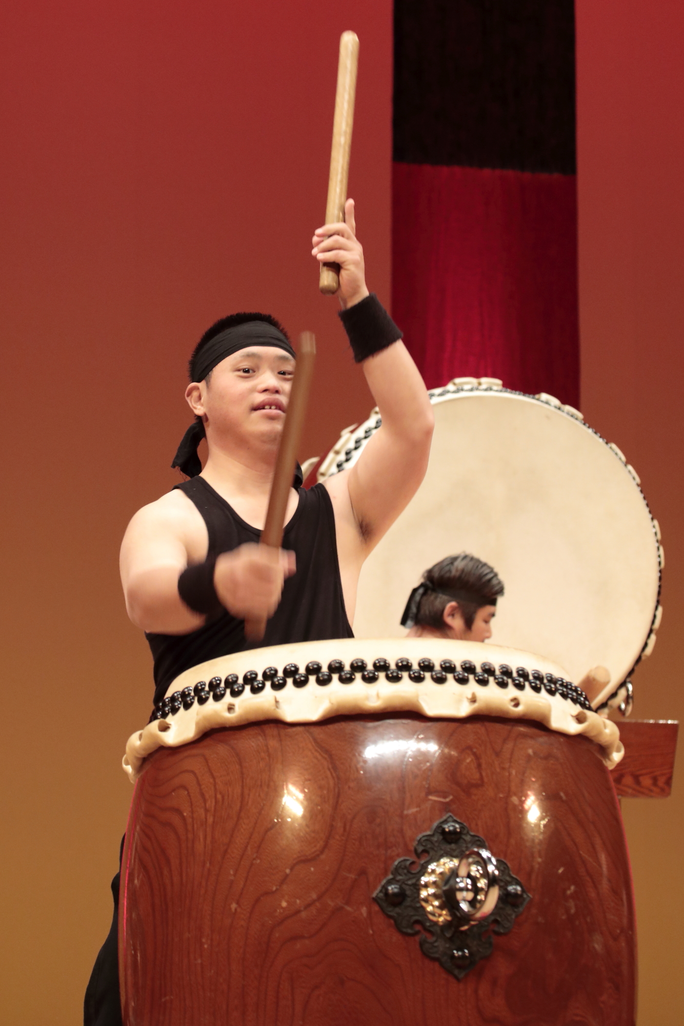 舞台の上で元気よくバチで和太鼓を叩く、黒い鉢巻を巻いた衣装姿の男性の写真