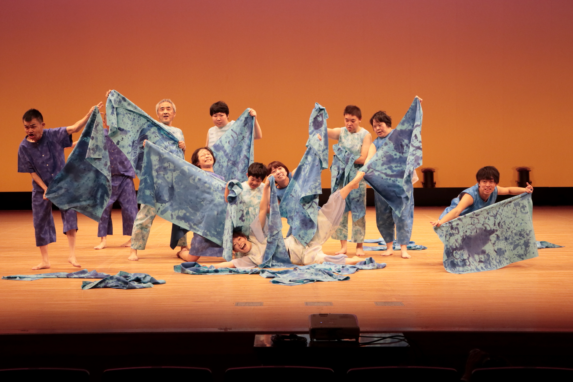 ライトが当たった舞台の上で、絞り染めの布を広げている人たちのダンスの様子の写真