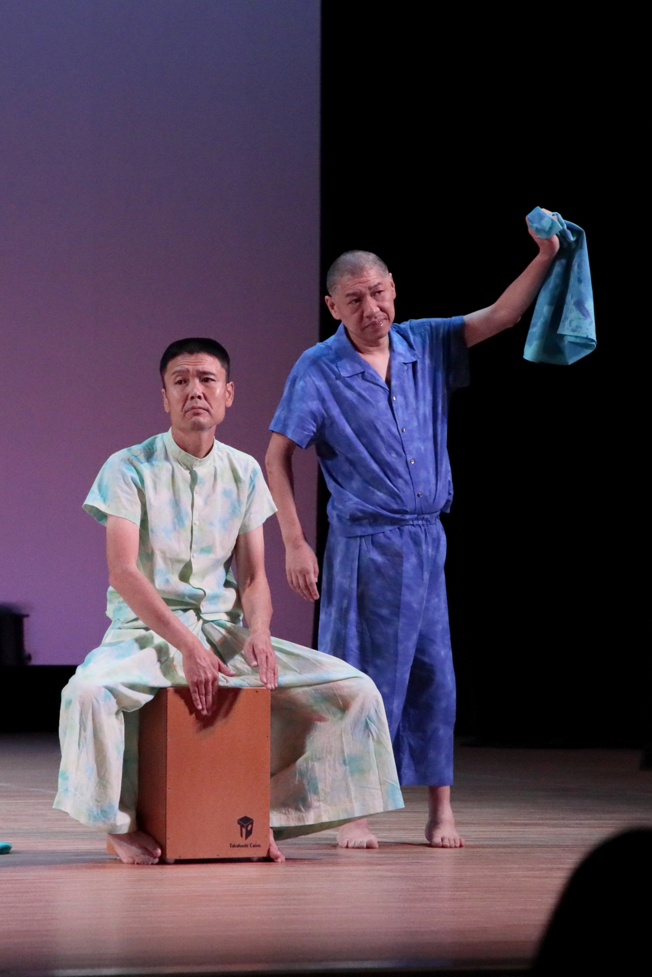 スポットライトが当たった舞台の上で、箱に跨ったり青い絞り染めの布を持ち上げている2人の男性の写真