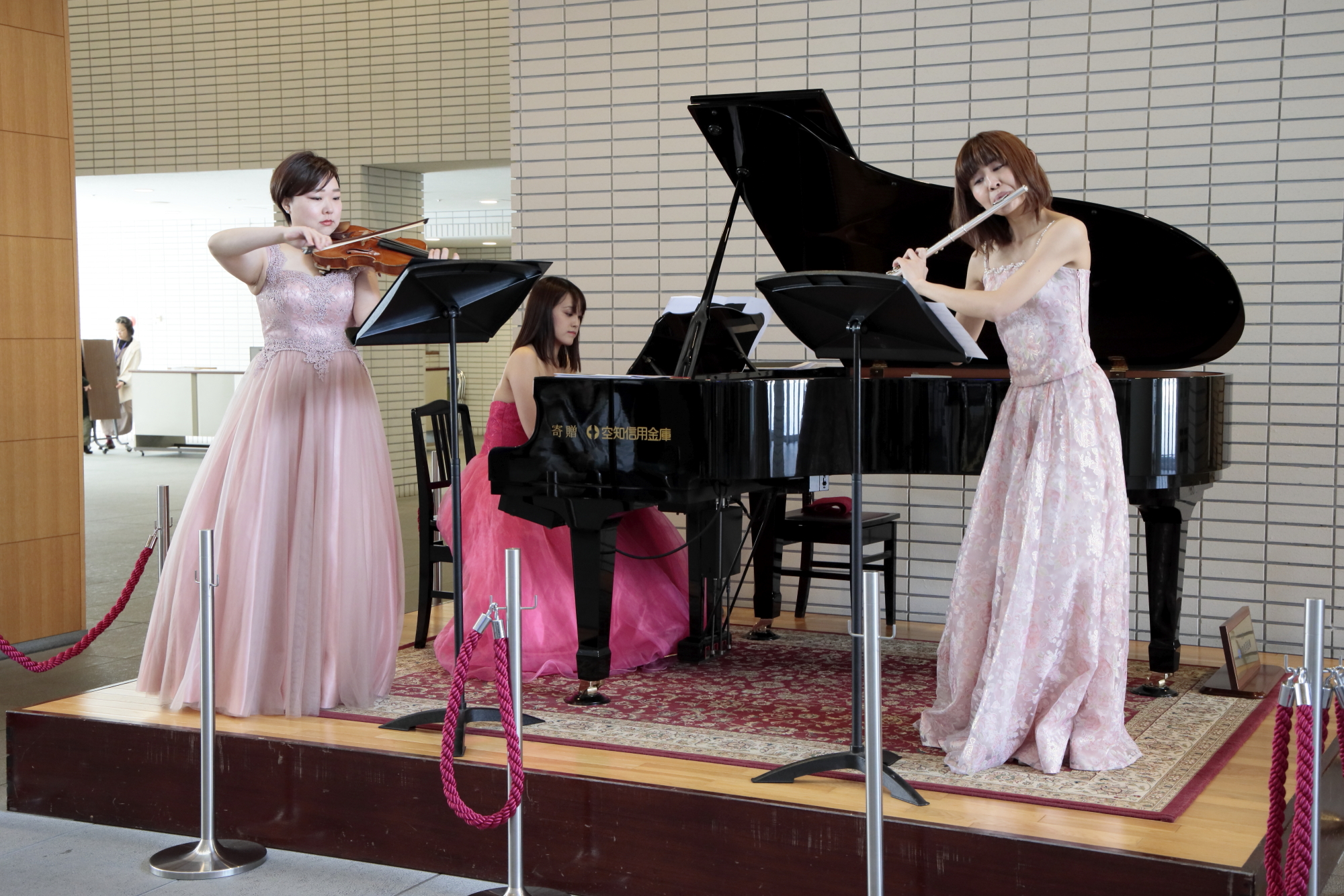 ロビーの小さな壇上でピアノとバイオリンとフルートを演奏する、ドレス姿の女性たちの写真