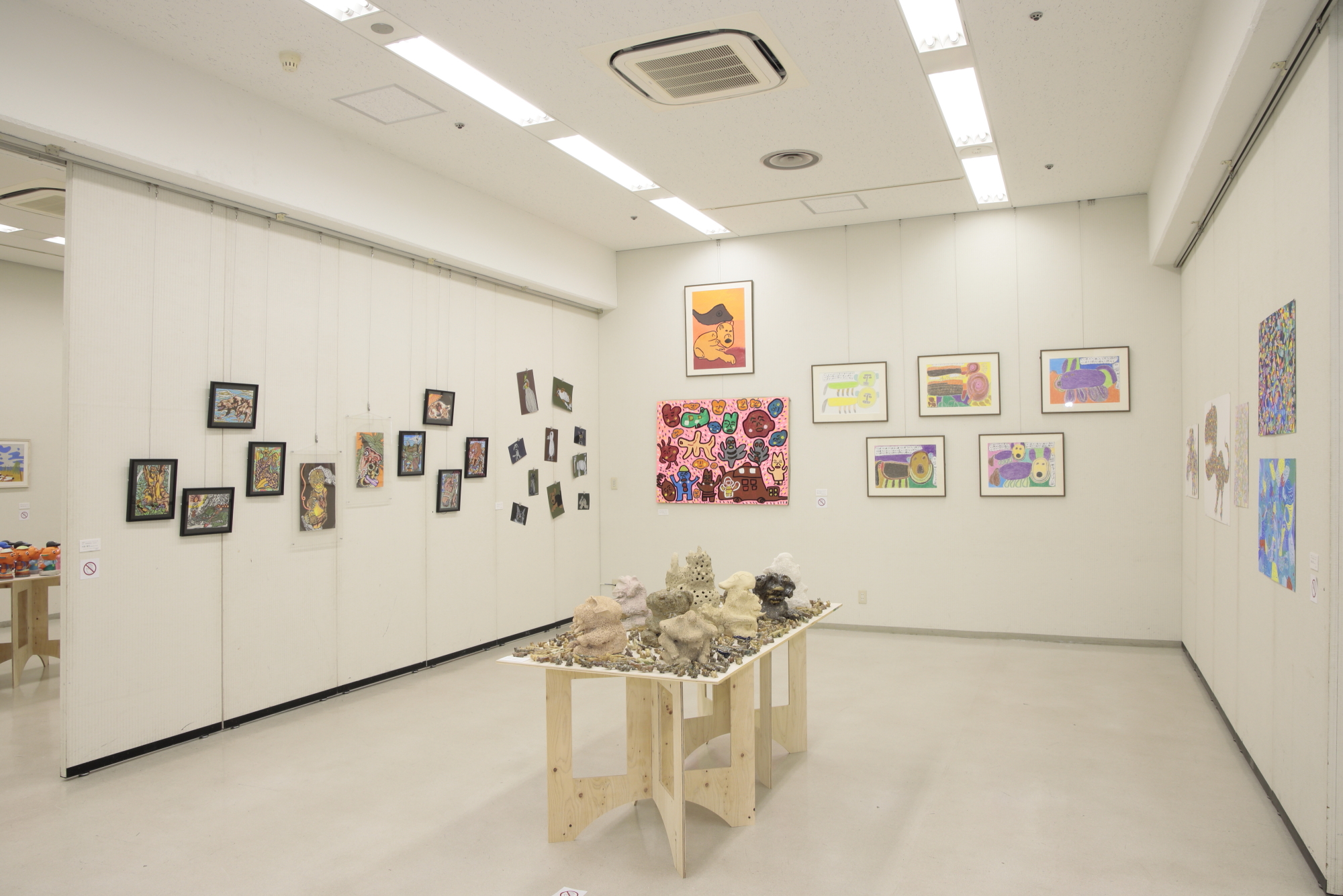 室内の白い壁に飾られた絵画や、中央のテーブルに作品が置かれた展示会の写真