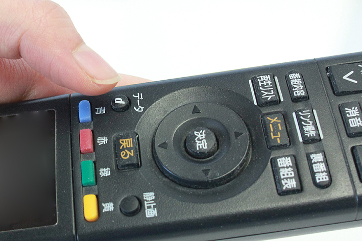 リモコンを左手に持ち、「dボタン」を押そうとする親指の写真