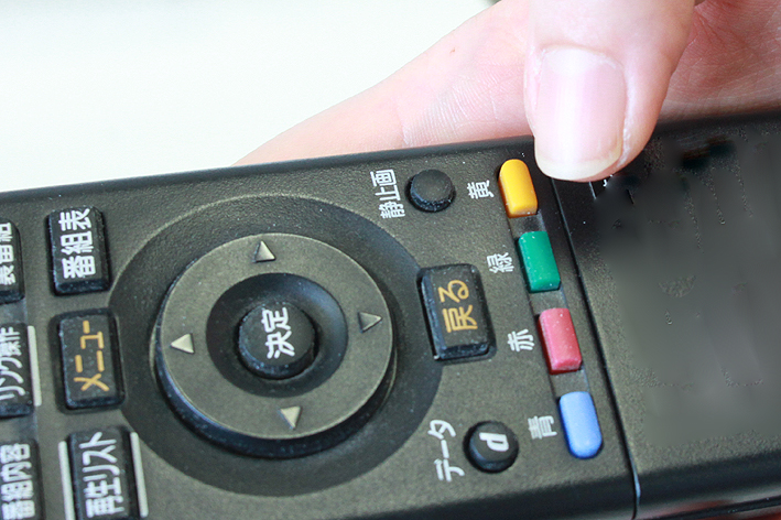 リモコンを右手に持ち、「黄色ボタン」を押そうとする親指の写真