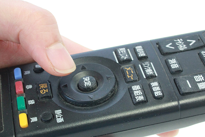 リモコンを左手に持ち、上下のボタンの下ボタンを押そうとする親指の写真