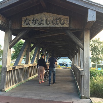 なかよしばしと書かれた木造の橋をベビーカーを押した女性と男性が並んで手前から奥に歩いている様子の写真