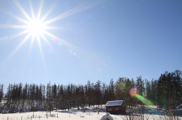 手前に小屋と木々の雪景色、背景に青空と太陽が写っている写真