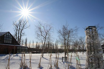 左側に三角屋根の建物と手前に雪景色、背景に青空と太陽が写っている写真