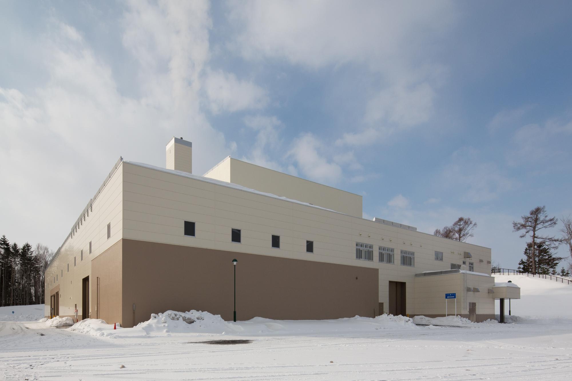 雪の中、クリーム色の壁面の四角い建物が立っている「いわみざわ環境クリーンプラザ」の施設外観写真