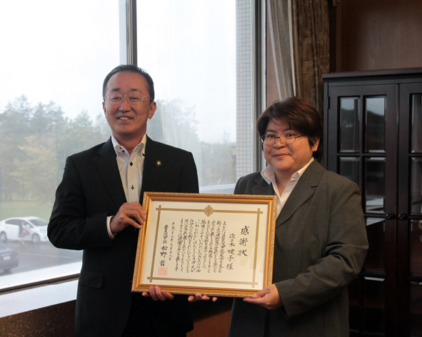 表彰状をカメラのほうへ向けて笑顔で並ぶ市長と佐々木さんの写真