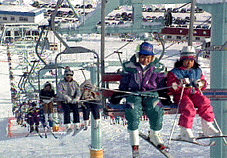 晴れた日のスキー場で、スキーを履きストックを手にした人達が、リフトに乗っている写真。二人掛けのリフトに二人ずつ、座っている。周りは白く雪がつもっている。