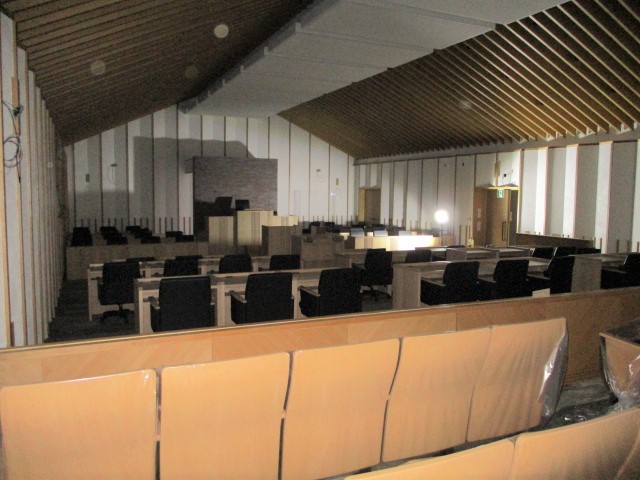 白い壁があり、椅子が中央に向けて並べられている議場の写真