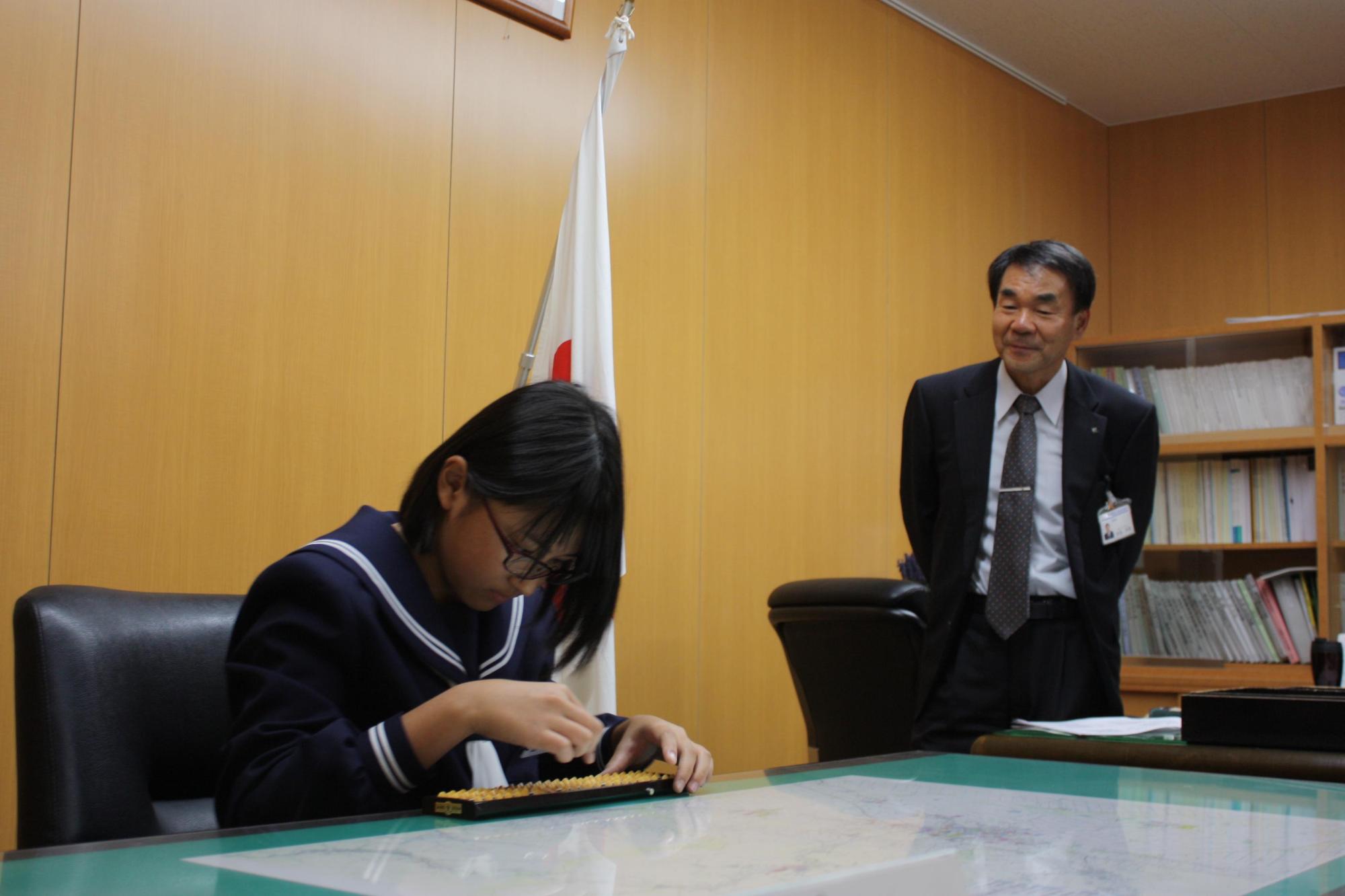 山元さんがそろばんをする様子を眺める教育長の写真