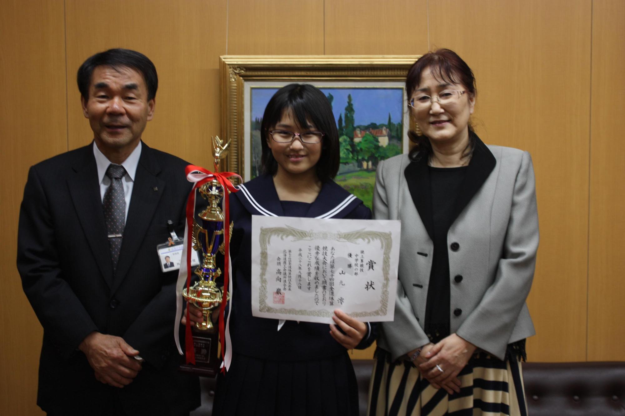 トロフィーと賞状を持つ山元さんと山元さんと女性と教育長が並ぶ写真