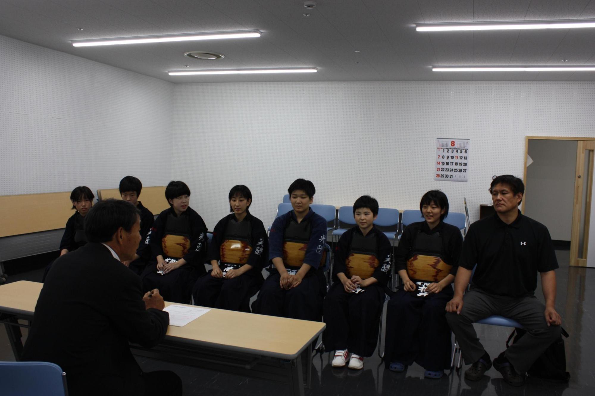 机を挟み教育長と生徒の皆さんと顧問が話を聞いている様子の写真