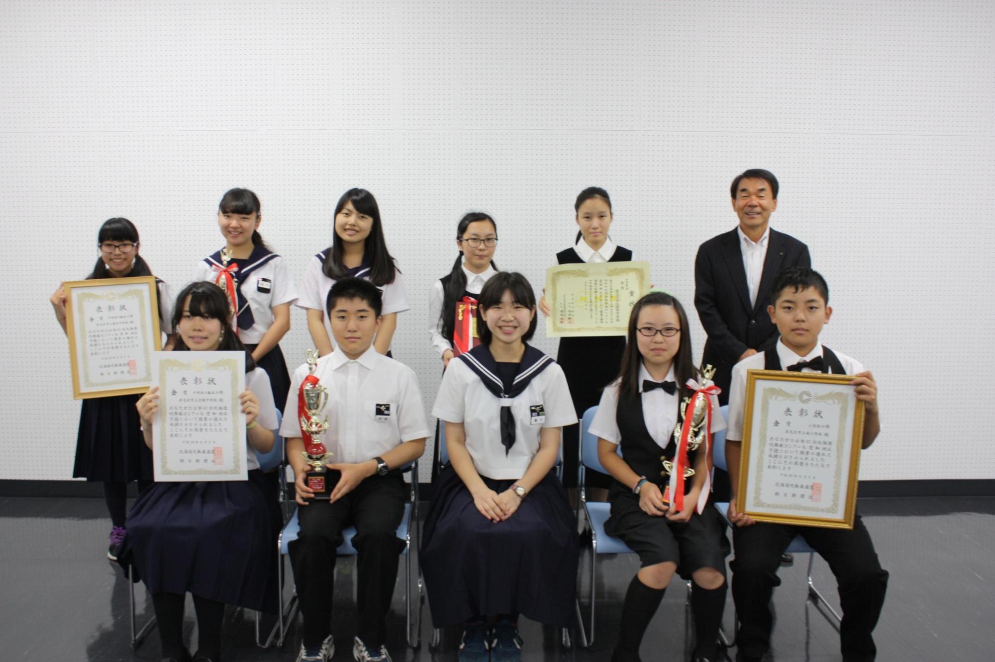 椅子に座ったりその後ろに立ち賞状トロフィーを掲げる吹奏楽部のみなさんと教育長の写真