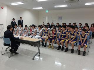 バスケットボールのユニフォームを着た子供数人と保護者が、机を挟んで教育長と話をしている写真。