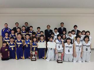 バスケットボールのユニフォームを着た子供数人,保護者と教育長が記念撮影をした写真。