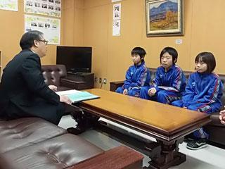 スーツを着た男性と机を挟んで、3人の青いユニフォームを着た生徒が座っている写真