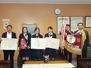 賞状、トロフィー、優勝旗と一緒に記念撮影する簿記部の女子生徒の三人と教育長