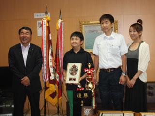 優勝旗とトロフィーを脇に立て盾を持って教育長と記念撮影する男子生徒の様子