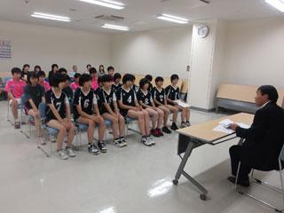バレーボールのユニフォームを着た女子数人とバドミントンのユニフォームを着た男子数人と保護者が、机を挟んで教育長と話をしている写真。