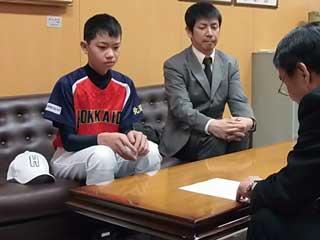 スーツを着た男性と机を挟んで、1人のスーツを着た男性と、赤と青のユニフォームを着た男子生徒が座っている写真