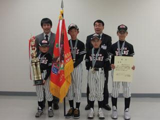 右から、賞状を持った男子生徒、手を後ろで組んでいる男子生徒、旗を持っている男子生徒、トロフィーを持っている男子生徒が並び、その後ろにスーツを着た男性が2人並んでいる写真