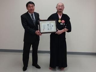 右の黒い道着を着た男性と、左のスーツを着た男性が2人で賞状を持って並んでいる写真