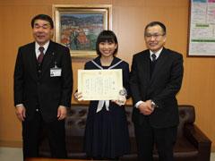 右から、スーツを着た男性、制服を着て賞状をこちらに向けている女子生徒、スーツを着た男性が並んで立っている写真