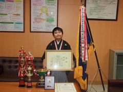机に沢山のトロフィーが飾られている応接室で、袴姿の少年が、優勝旗の隣で賞状を持っている写真