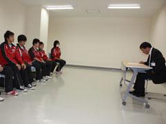 白い壁の明るい室内で、ジャージ姿の学生数名が、向かいに座っている男性と話をしている写真
