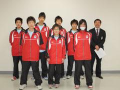 白い壁の明るい室内で、赤いジャージ姿の学生7名と男性1名が、2列に並んで立っている記念写真