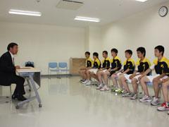 右手に黄色と黒と白のユニフォームを着た選手たちが並んで座っていて、右手にスーツ姿の男性が座っている写真
