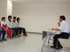 左手に選手が4人椅子に座って横並びになっていて、右手に男性が机の前の椅子に座っている写真