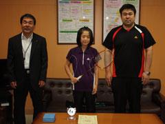真ん中にバトミントンを持ち紫色のユニフォームを着て立っている女子学生がいて、その両隣に男性が立っている写真