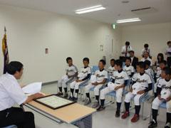 奥に白いユニフォームを着た選手たちが沢山座っていて、手前に男性が机の前の椅子に座っている写真