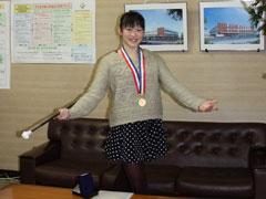 応接室のソファーの前で、メダルを首から下げた女生徒が、立ってバトンを持ちながらポーズを決めている写真