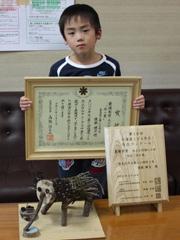 作品と一緒に表彰状を持ちながらたつ児童の写真