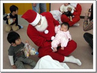 サンタクロースの膝の上で眠る乳幼児を隣でプレゼントの袋を握った男の子が見つめている写真