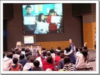 長崎県五島市立緑丘小の児童3名が映る大きなスクリーンに向かい身を乗り出し手をあげる児童たちの写真