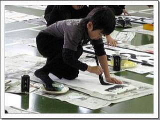 床に広げた新聞紙の上に半紙を置き大きな筆で書初めをする男子児童の写真