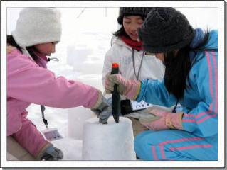 雪野原の上で形よく固めた雪をいくつも並べ、スコップで穴を掘る3人の女子児童の写真