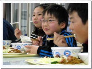 ご飯のどんぶりを手に緑のトレーに乗ったおかずをほおばり笑顔の児童たちのアップの写真