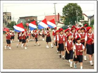 赤白青の旗や楽器を持って学校のグラウンドで行進をしている、赤いベレー帽をかぶった制服姿の生徒たちの写真