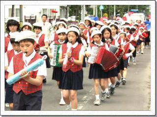 楽器を演奏しながら町中を行進する、白いベレー帽をかぶった制服姿の生徒たちの写真