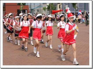 町中で指揮棒を振りながら、楽器を持った多くの生徒たちを先導している女子生徒の写真