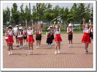 町の広場で整列をして楽器を演奏している生徒たちの前で、バトンを頭上に放っている制服姿の女子生徒たちの写真