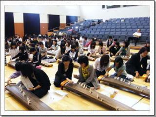 講堂で大勢の大学生が小学生たちに寄り添い、琴を教えている写真