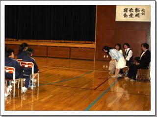 体育館の椅子に座った生徒に向けて、起立して頭を下げている女性の写真