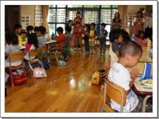 幼稚園でお弁当が乗った机に向かい手を合わせる子供たちと、みんなの前で手を合わせる先生の写真